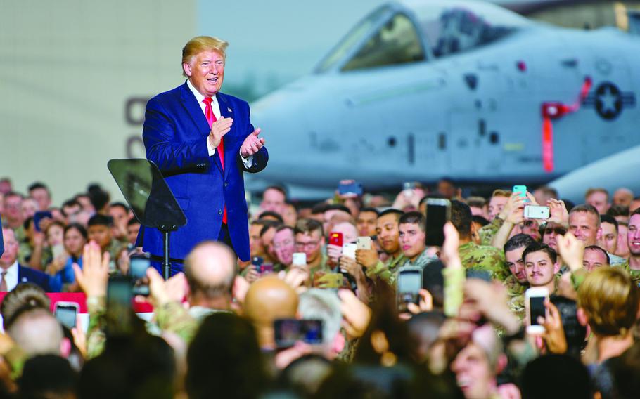 Donald Trump stands on a platform surrounded by service members and their families, many holding up phones, in front of a mlitary aircraft at Osan Air Base.