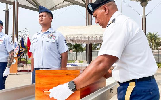 A soldier in dress uniform and white gloves places a wooden case into a casket, while another soldier in dress uniform stands at attention next to the casket.