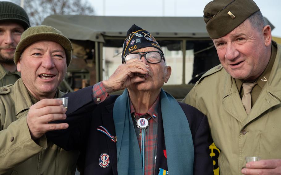 101-year-old WWII veteran Ceo Bauer drinks from a shot glass while World War II reenactors on either side of him hold shot glasses for the camera.