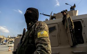 A masked man with a rifle and yellow arm patch is seen in the foreground with a tank in the background.