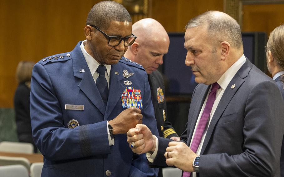 A man in uniform wearing glasses and standing touches his fist to the fist of another man who is also standing and wearing a gray suit. 