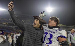 A man in a pirate hat takes a selfie with a football player on a football field.