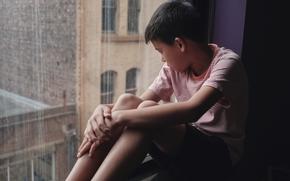 A young boy gazes thoughtfully out of a rain-streaked window. 