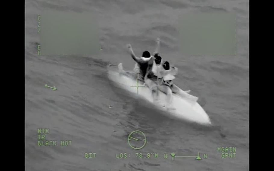 Three boaters wave from the hull of a capsized boat