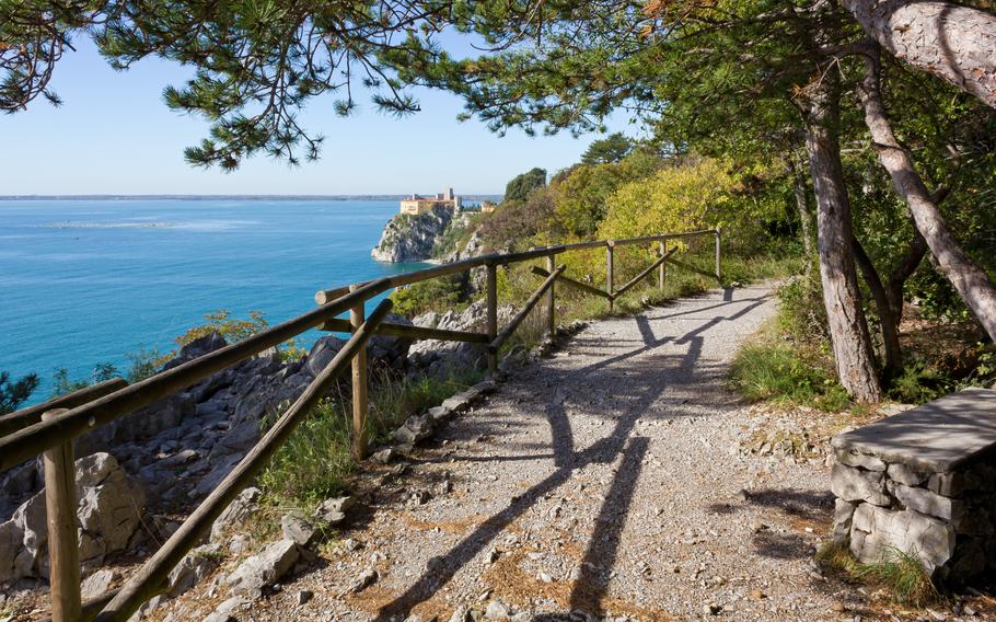 The ocean extends beyond a dirt trail with a wooden fence on one side and trees on the other. A cliffside stone structure is visible in the far distance.