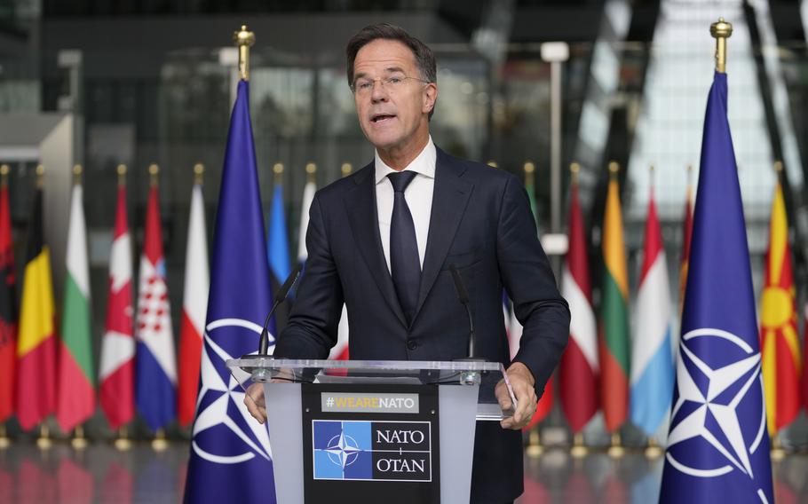 NATO Secretary General Mark Rutte wears a suit and stands in front of flags of many nations.