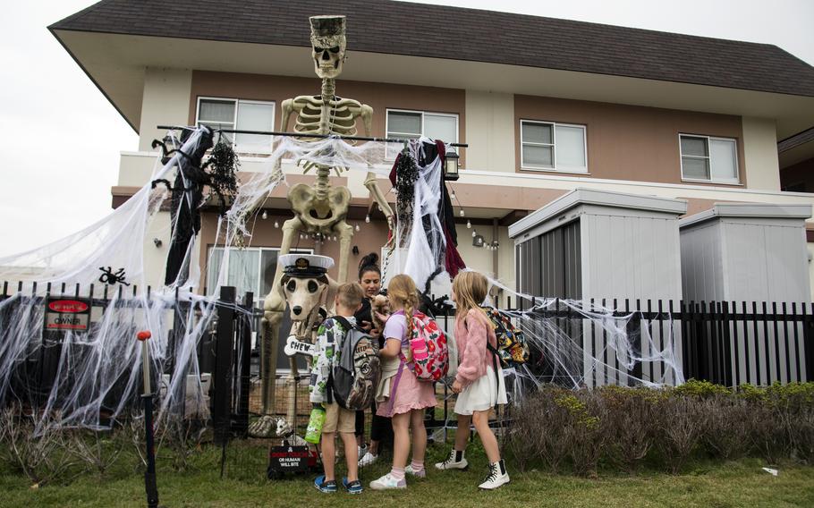 A group of children gather by a fence next to a large skeleton Halloween decoration in front of a house.