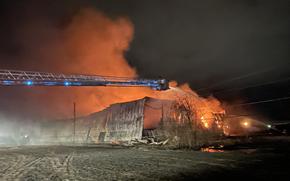 Night view of firefighters in the bucket of a fire truck ladder using a hose hose to spray water on the flames from a fire in a storage facility.