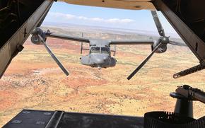 A view from the interior of one Osprey shows a second Osprey behind it with the orange and yellow landscape below dotted with green.