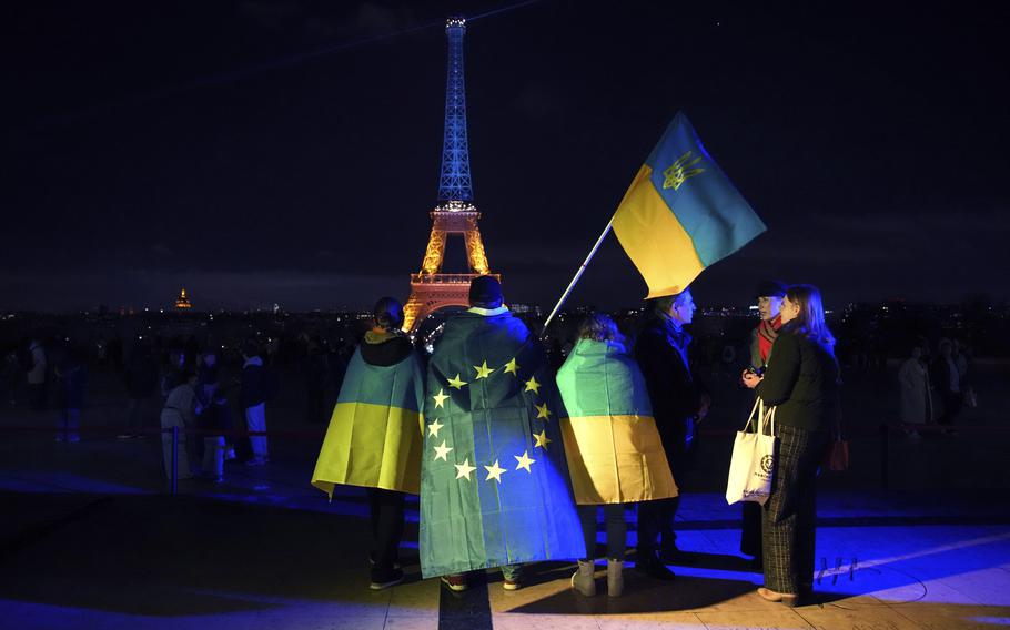 People in front of the Eiffel Tower at night, two wearing Ukrainian flag coats and one carrying the Ukrainian flag.