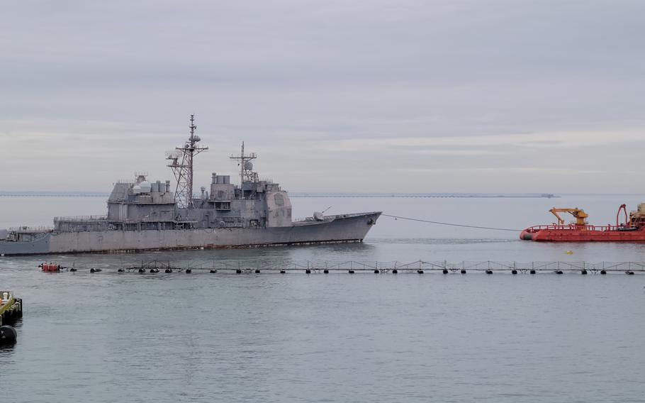 A Navy cruiser on the left is seen being towed by a smaller orange ship on the right.