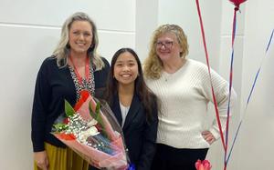 A girl holding a bouquet of red and white flowers stands smiling with her school’s principal and vice principal.