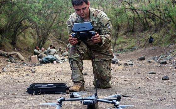 A soldier looks at a digital display screen while kneeling on the ground next to an unmanned aerial vehicle.