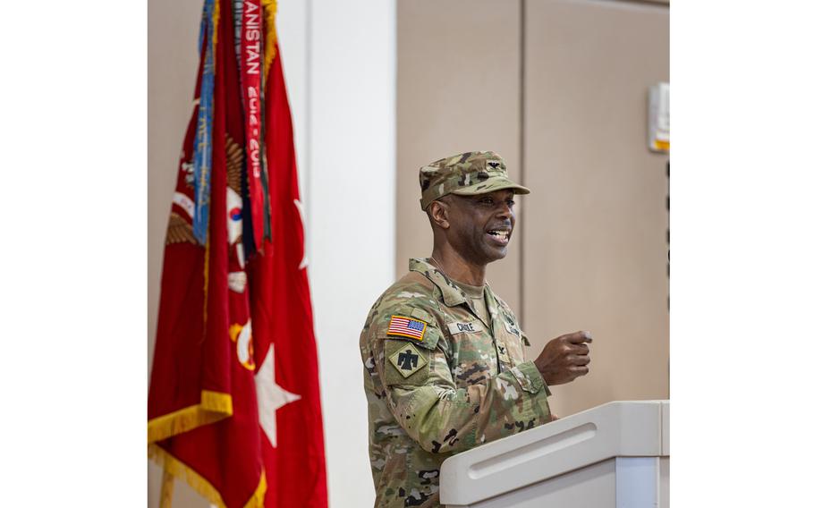 An Army colonel speaks at a podium with one hand raised in gesture, next to flags.