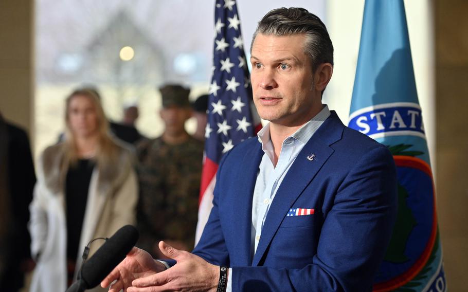 Pete Hegseth, wearing a blue blazer, speaks into a microphone while standing in front of a U.S. flag.