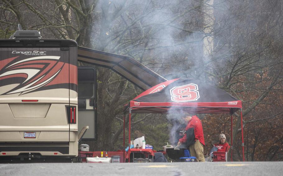 A man stands under a red tent and grills in a parking lot.