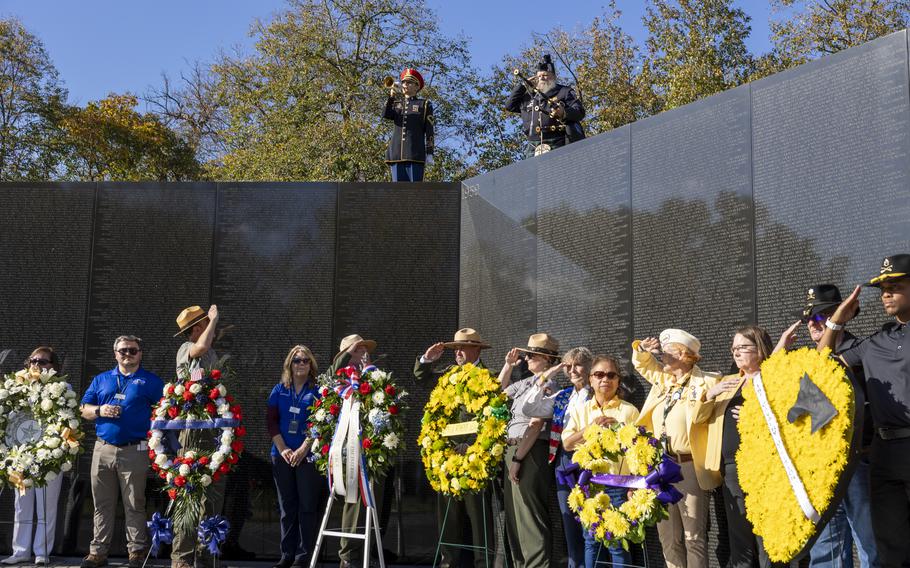 A military member plays the trumpet from above the veterans memorial while people in front of the memorial salute.