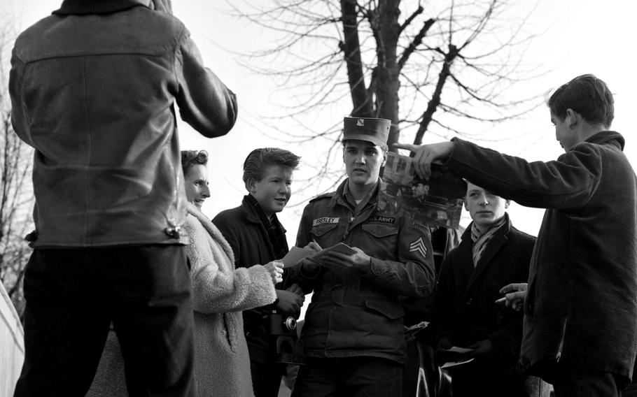 Elvis Presley wears his Army uniform while signing autographs, surrounded by fans.