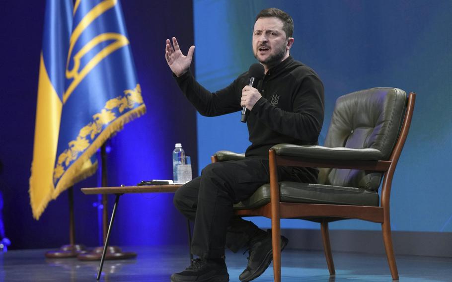 Zelenskyy holds a microphone in his left and raises his right hand as he speaks.