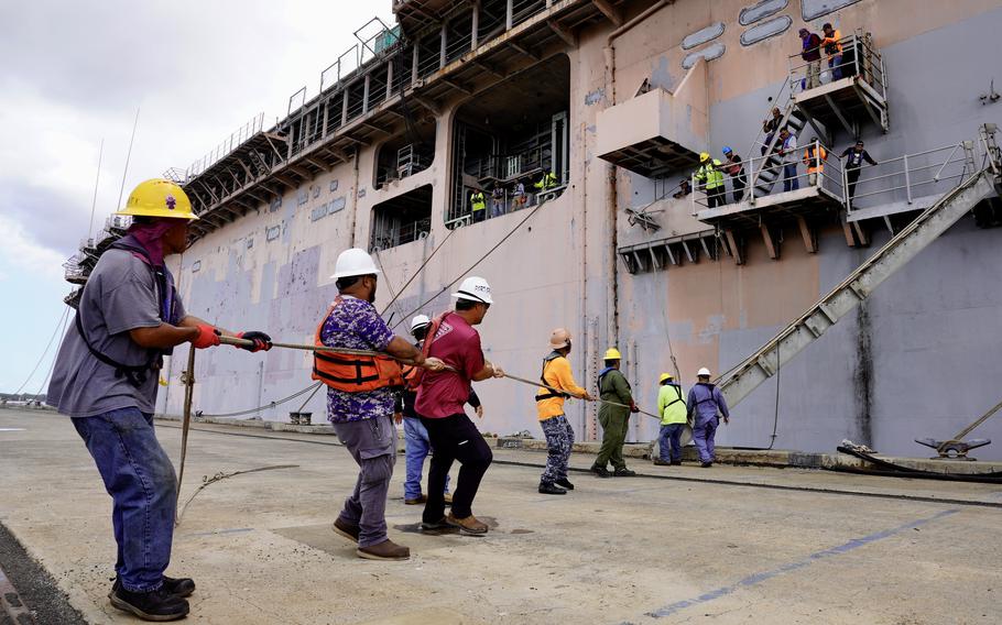 Shipyard workers line up to pull a rope attached to a ship’s hull.