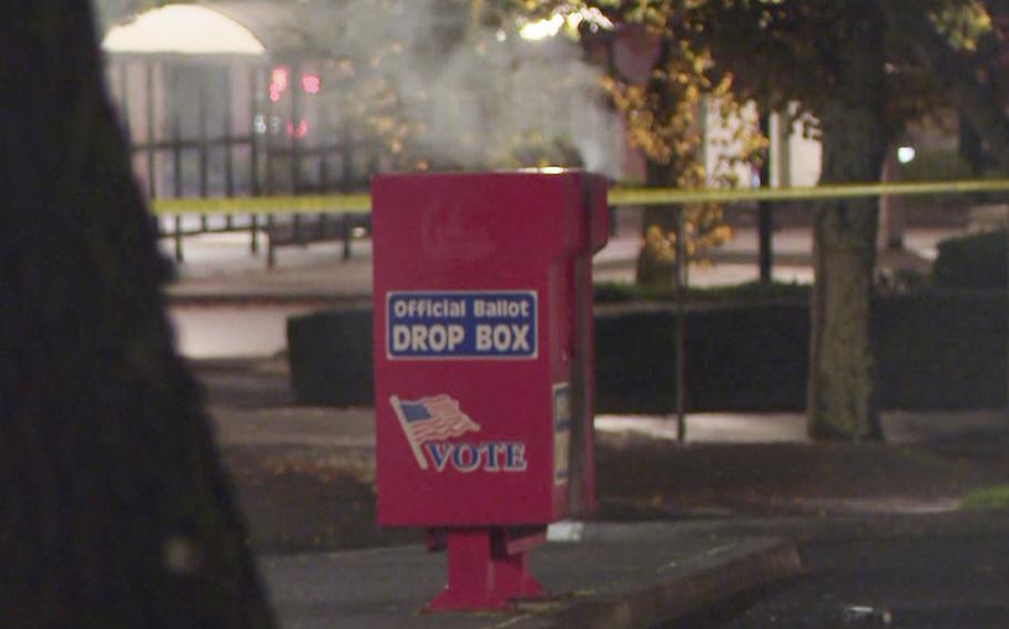 Smoke pours out of the top of a red ballot drop box on a sidewalk.