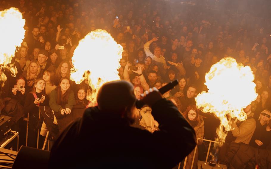 A man wearing a hoodie holds a microphone and sings in front of a crowd on a stage outfitted with pyrotechnics.