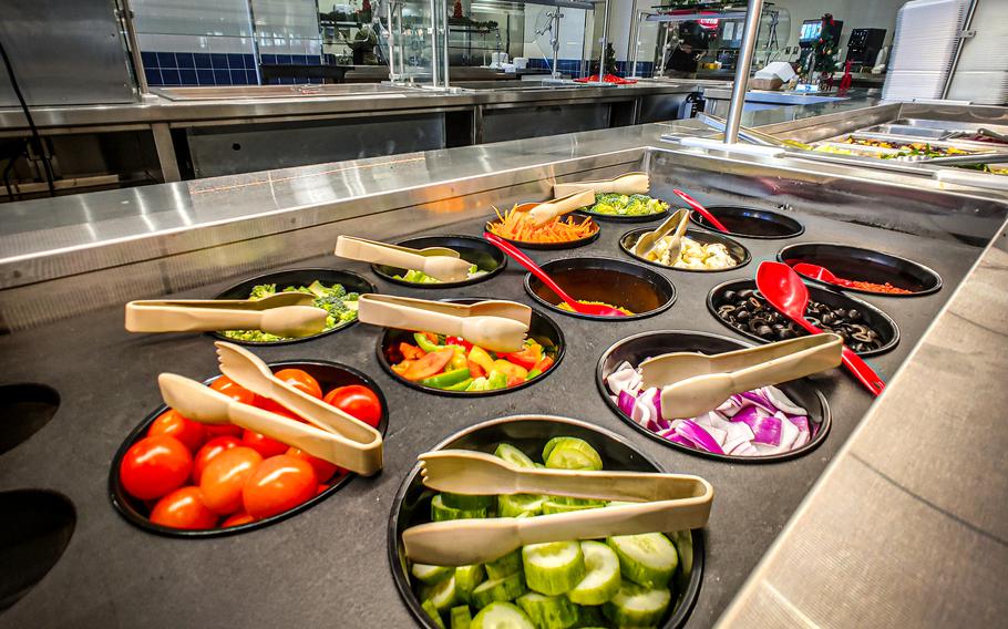 Tongs and spoons sit inside containers on a salad bar with tomatoes, cucumbers, purple onions, bell pepper slices, olives and more.