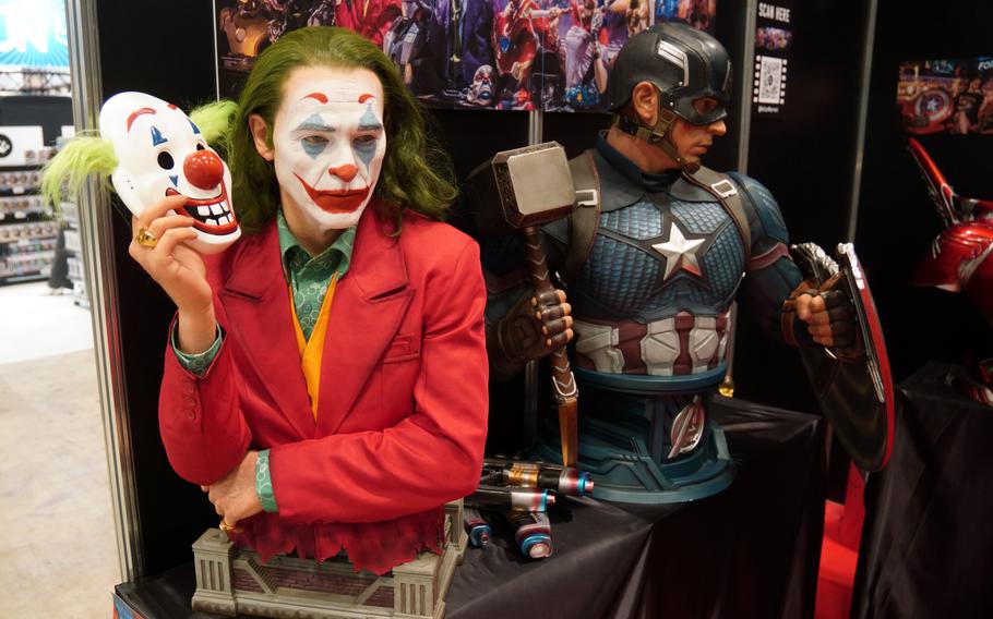 A bust of a man with green hair wearing a red suit, clown makeup and holding a clown mask is displayed on a table.