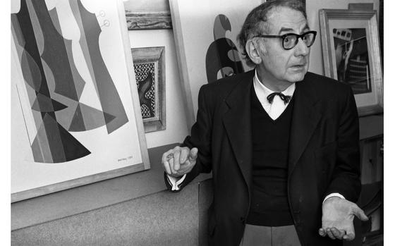 Paris, France, Sep. 22, 1965: Artist Man Ray - photographed in his Paris studio - stands before some of his works. To his right stands a print of "Orchestra," one of the 10 pochoirs (stencils) from the series titled "Revolving Doors." Right behind him is a print titled "Shadows," from the same series. Man Ray produced the original "Revolving Doors" series as collages between 1916 and 1917. 
The collages, whose geometric shapes combine machine-like and anthropomorphic forms, were framed and installed on a rotating pole that the viewer could spin. The original collages were destroyed, but Ray later reproduced them as limited edition stencil prints (pochoirs), published by Éditions Surréalistes in Paris.

META TAGS: art; culture; artist; painter; photographer; Dada; dadaism; surrealism; surrealist; film maker; stenciling; pochoir; stencil; avant-garde; Emmanuel Radnitzky