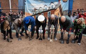 Seven people lean forward holding shovels to scoop dirt to mark the groundbreaking for the Tun Tavern.