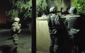 A soldier and interpreter, both in combat gear and seen from behind, look over a stone wall during a night patrol.