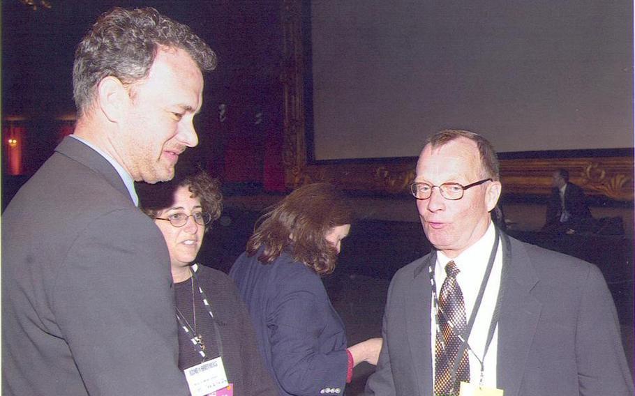 Actor Tom Hanks stands at the left with his side to the camera as David Stewart stands at right talking.