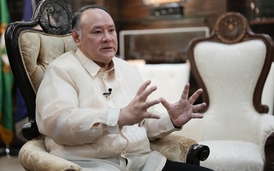 A man in a white collared shirt gestures with both hands while seated in a sofa chair.