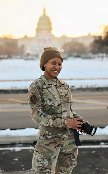 An airman with a camera flashes a smile with the Capitol in the background.