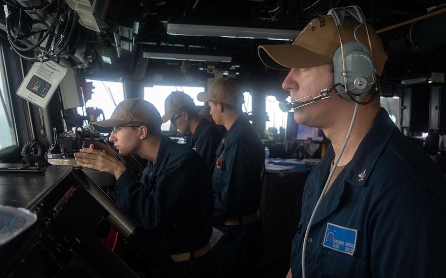 Men in khaki ball caps and navy uniforms look out from the pilot house of a ship.