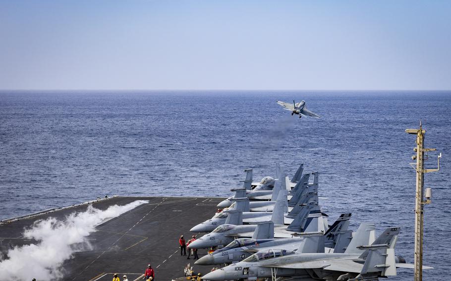 The flight deck of the carrier USS Harry S. Truman shows planes lined up, crew members performing their duties and a fighter jet taking off.