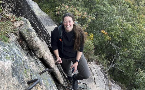 A woman smiles as she stands on a hiking or climbing trail.