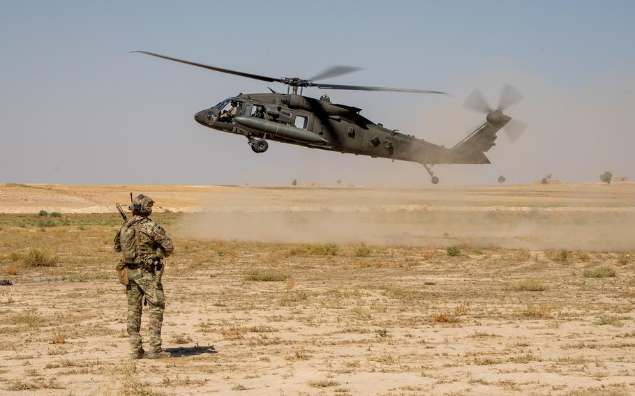 A Black Hawk helicopter kicks up dust as it lands in desert terrain in front of a U.S. service member in combat gear.