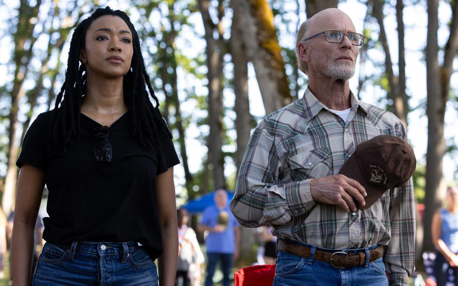 A young woman stands next to an older man, with the man holding his hat to his heart.