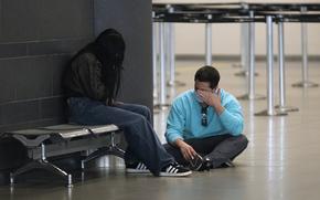 A woman sits on a seat with her face obscured by hair while a man sits cross-legged next to her on the floor, wiping his eyes.