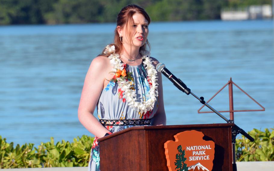 A woman wearing a floral dress and a flower lei speaks at the podium with an ocean view behind her.