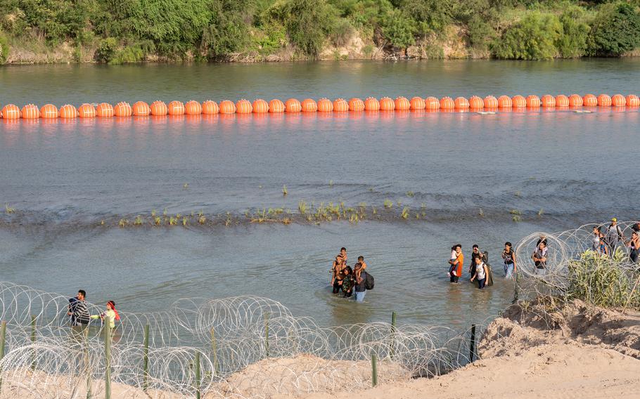 Migrants walk by a string of buoys placed on the water along the Rio Grande border with Mexico in Eagle Pass, Texas, on July 16, 2023. The buoy installation is part of an operation Texas is pursuing to secure its borders, but activists and some legislators say Texas Gov. Greg Abbott is exceeding his authority. 