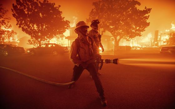 Firefighters in a burning neighbordhood spray a water hose at the flames.