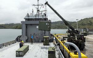 A crane on a pier lifts a cargo container onto the deck of a docked landing craft.