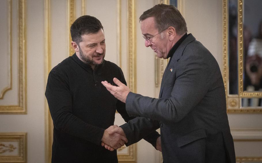 Volodymyr Zelenskyy, left, shakes hands with Boris Pistorius, who is smiling and raising his other hand in greeting.