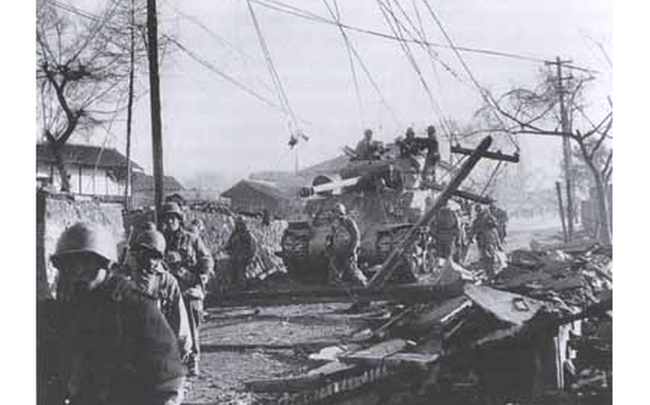 A black and white photograph of a tank rolling through wreckage.