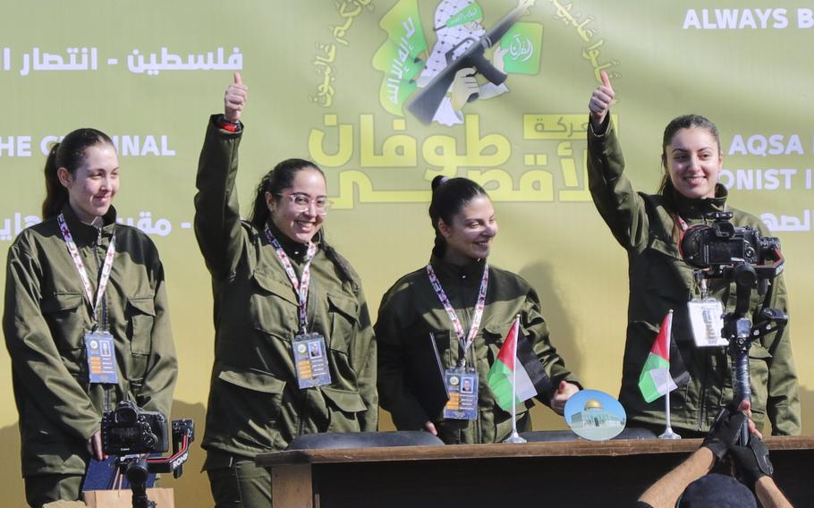 Four women dressed in army green clothing with lanyards around their necks smile and give thumps-up while standing on a stage.