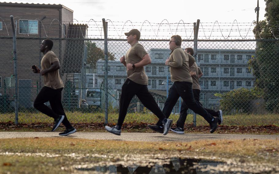 Runners jog past a chainlink fence next to a grassy patch marked with puddles during a 5K race.