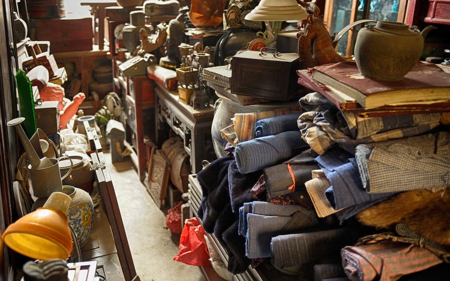 Many items such as rolled-up carpets, lamps and scrapbooks are stacked high in dusty piles.