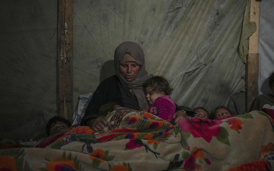 A woman in dirty clothing snuggles many young children that are tucked under a blanket.